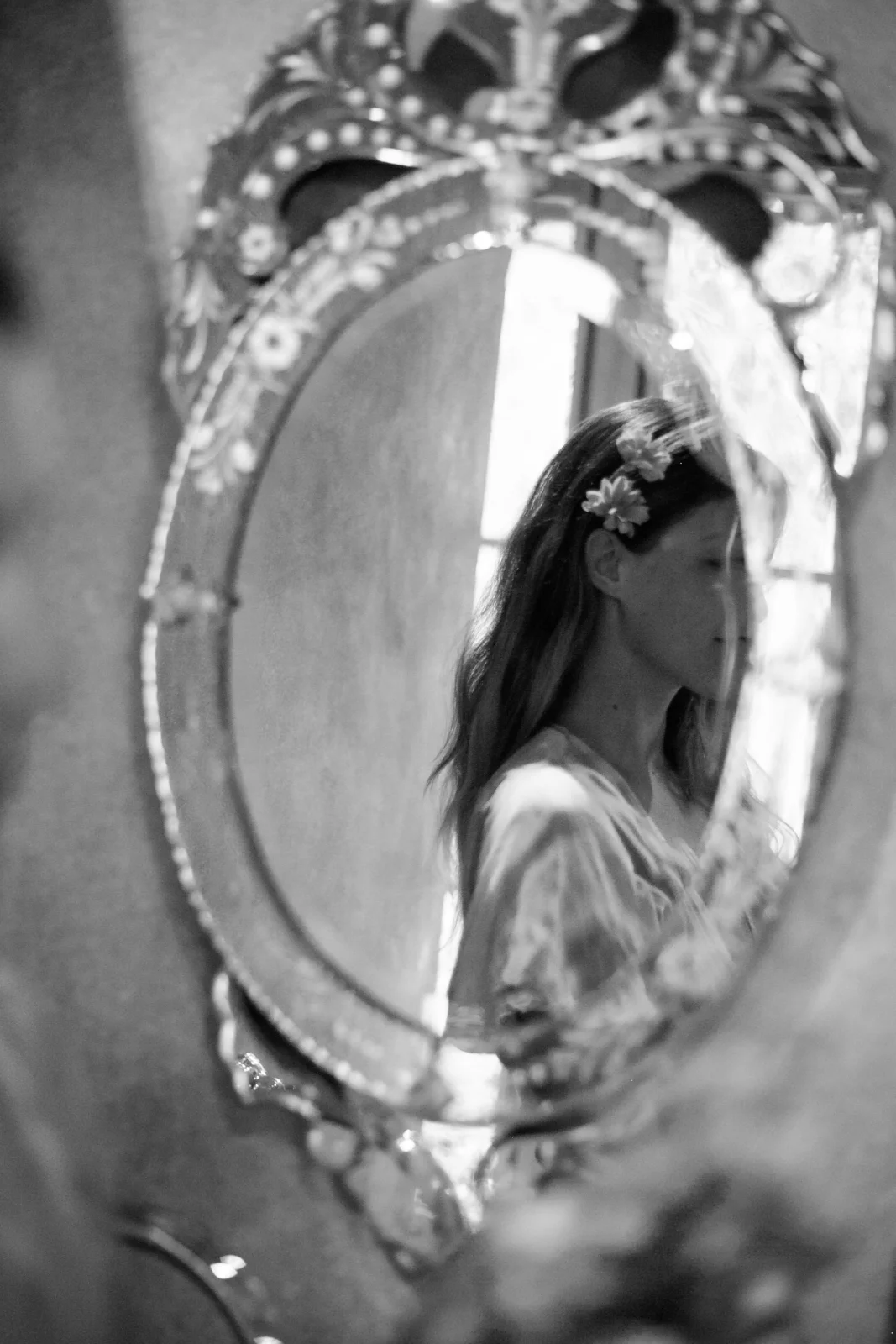 Photographie en noir et blanc d'une femme aux cheveux longs vue de profil, dont le reflet apparaît dans un miroir ovale au cadre richement orné. Elle porte de petites fleurs délicates dans ses cheveux. L'image possède une esthétique douce, ancienne et artistique.